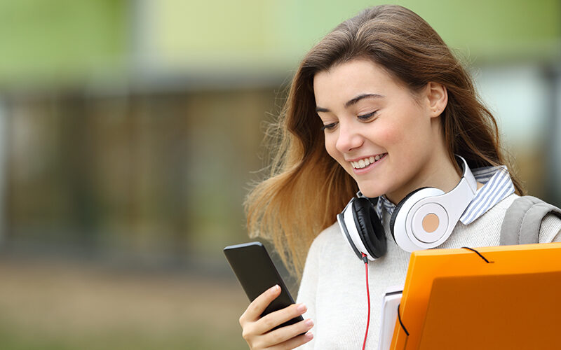 Estudante sorridente olha para o celular com fones de ouvido e pastas.