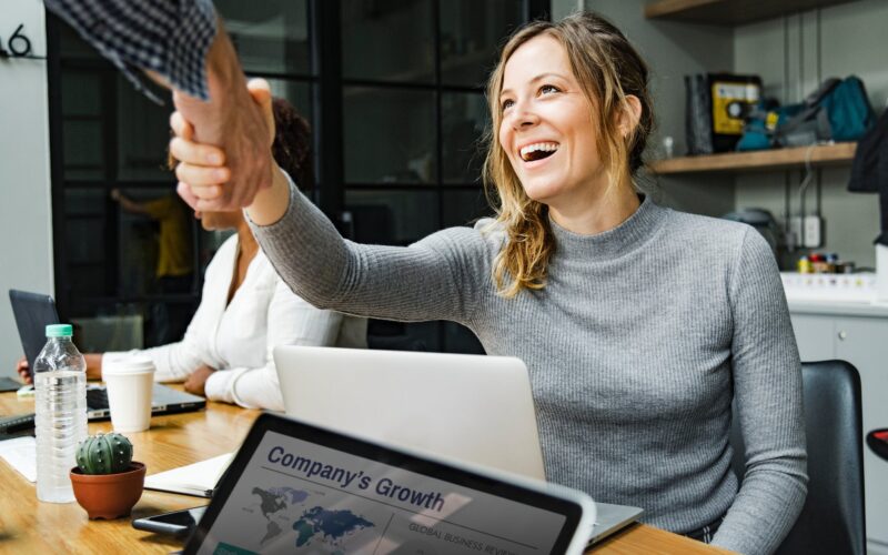 Uma jovem sorridente, de blusa cinza e cabelo loiro, aperta a mão de alguém, celebrando o seu primeiro emprego. Um laptop com está à sua frente, em uma mesa de escritório com garrafa de água, café e cacto.