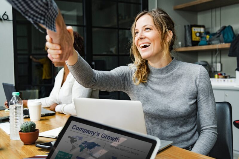 Uma jovem sorridente, de blusa cinza e cabelo loiro, aperta a mão de alguém, celebrando o seu primeiro emprego. Um laptop com está à sua frente, em uma mesa de escritório com garrafa de água, café e cacto.