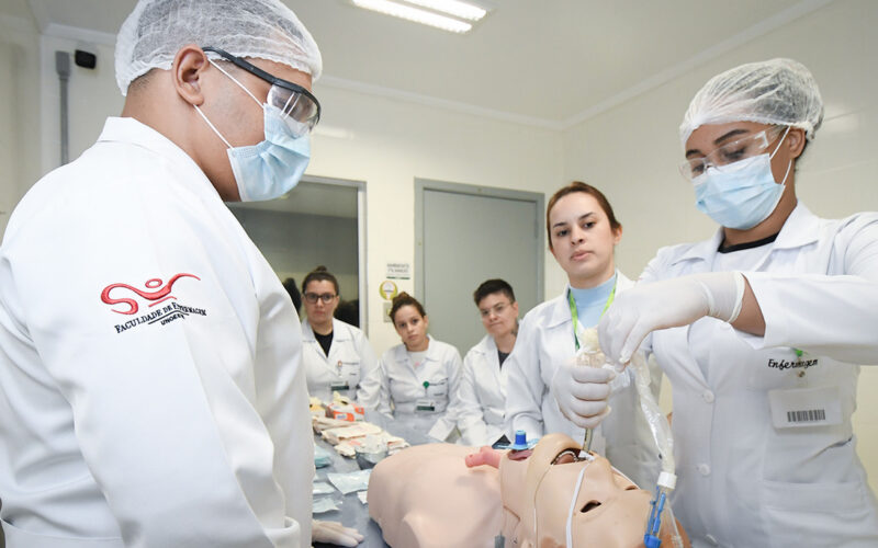 Estudantes do curso de Enfermagem da Unoeste participam de uma aula prática no Laboratório de Habilidades e Simulação, realizando procedimento de intubação em manequim de alta fidelidade sob supervisão, com equipamentos e materiais de suporte clínico dispostos na bancada.