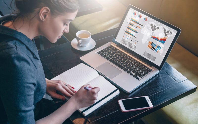 Mulher analisa gráficos no laptop e anota em caderno, com café e celular.