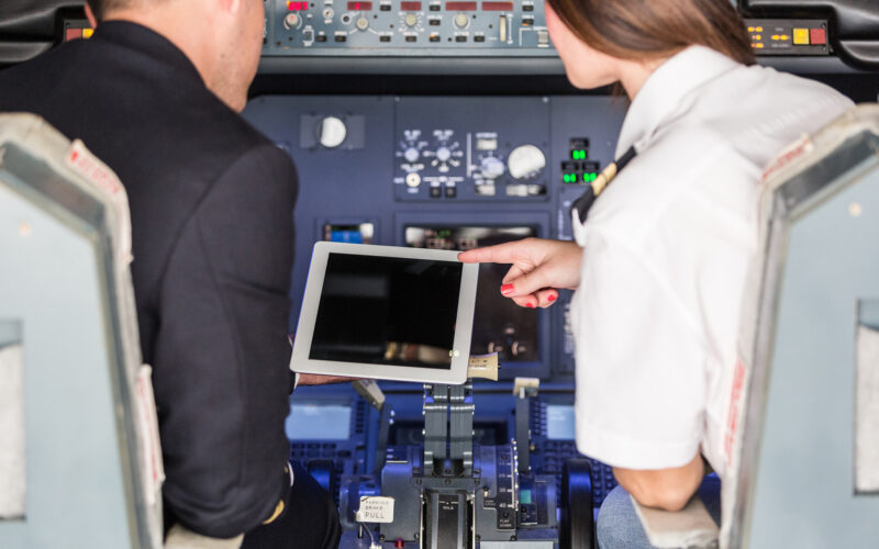 Pilotos usam tablet no cockpit do avião, painel de controle detalhado ao fundo.