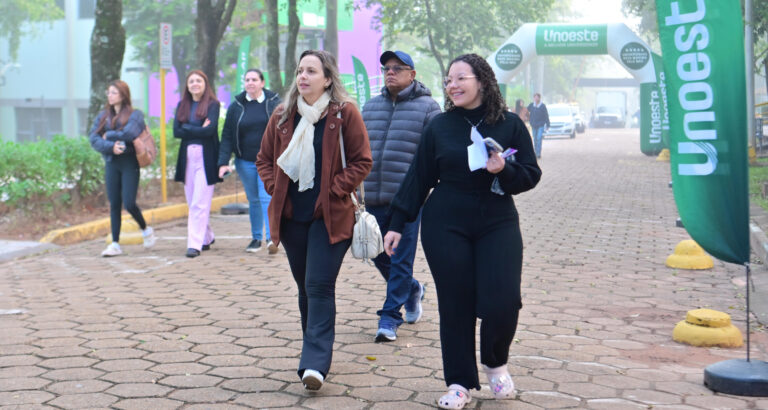 Pessoas caminham por uma área externa do campus da Unoeste, próximo à entrada sinalizada com bandeiras e arco institucional da universidade.