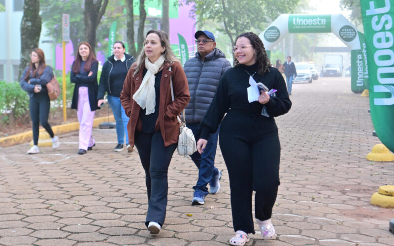 Pessoas caminham por uma área externa do campus da Unoeste, próximo à entrada sinalizada com bandeiras e arco institucional da universidade.