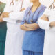 Um grupo diversificado de profissionais de saúde, incluindo médicos e enfermeiros, posa com os braços cruzados, vestindo jalecos brancos e uniformes azuis e verdes, com estetoscópios ao redor do pescoço
