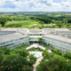 Vista aérea do campus da Unoeste em Presidente Prudente, São Paulo. Vários prédios de poucos andares, de cor clara, estão dispostos em torno de uma área central ajardinada, interligados por passarelas. A vegetação abundante ao redor do campus contrasta com a paisagem rural visível ao fundo.