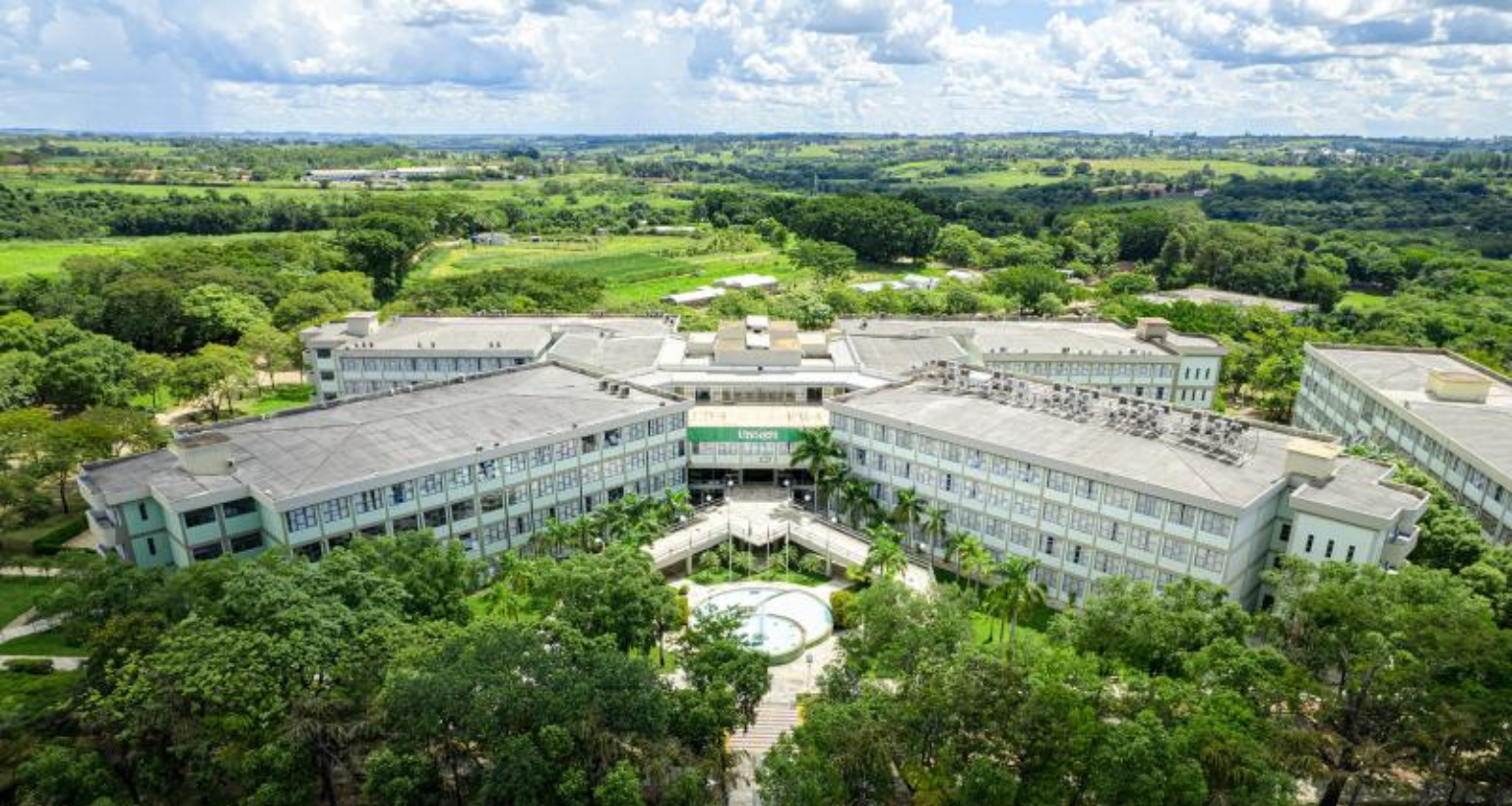 Vista aérea do campus da Unoeste em Presidente Prudente, São Paulo. Vários prédios de poucos andares, de cor clara, estão dispostos em torno de uma área central ajardinada, interligados por passarelas. A vegetação abundante ao redor do campus contrasta com a paisagem rural visível ao fundo.