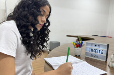 Estudante com cabelos cacheados escreve em caderno durante momento de estudo em sala da Unoeste, com objetos decorativos e letreiro com o nome da universidade sobre a mesa.