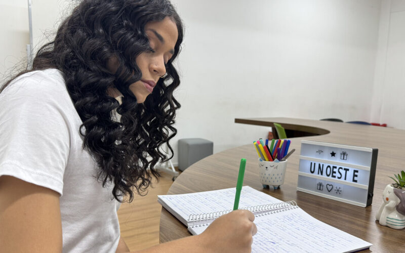 Estudante com cabelos cacheados escreve em caderno durante momento de estudo em sala da Unoeste, com objetos decorativos e letreiro com o nome da universidade sobre a mesa.