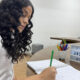Estudante com cabelos cacheados escreve em caderno durante momento de estudo em sala da Unoeste, com objetos decorativos e letreiro com o nome da universidade sobre a mesa.