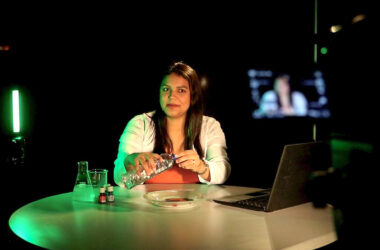 Uma mulher aparece sentada atrás de uma mesa redonda em um estúdio de gravação de videoaulas com iluminação verde ao fundo, demonstrando um experimento prático ao segurar uma garrafa plástica sobre um prato. À sua direita está um notebook aberto e, à esquerda, frascos de laboratório béquer e erlenmeyer, enquanto a silhueta de uma câmera de filmagem em primeiro plano e um monitor ao fundo contextualizam a produção de conteúdo educacional EAD da Unoeste.