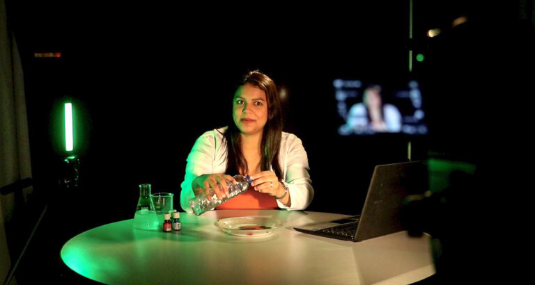 Uma mulher aparece sentada atrás de uma mesa redonda em um estúdio de gravação de videoaulas com iluminação verde ao fundo, demonstrando um experimento prático ao segurar uma garrafa plástica sobre um prato. À sua direita está um notebook aberto e, à esquerda, frascos de laboratório béquer e erlenmeyer, enquanto a silhueta de uma câmera de filmagem em primeiro plano e um monitor ao fundo contextualizam a produção de conteúdo educacional EAD da Unoeste.