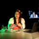 Uma mulher aparece sentada atrás de uma mesa redonda em um estúdio de gravação de videoaulas com iluminação verde ao fundo, demonstrando um experimento prático ao segurar uma garrafa plástica sobre um prato. À sua direita está um notebook aberto e, à esquerda, frascos de laboratório béquer e erlenmeyer, enquanto a silhueta de uma câmera de filmagem em primeiro plano e um monitor ao fundo contextualizam a produção de conteúdo educacional EAD da Unoeste.