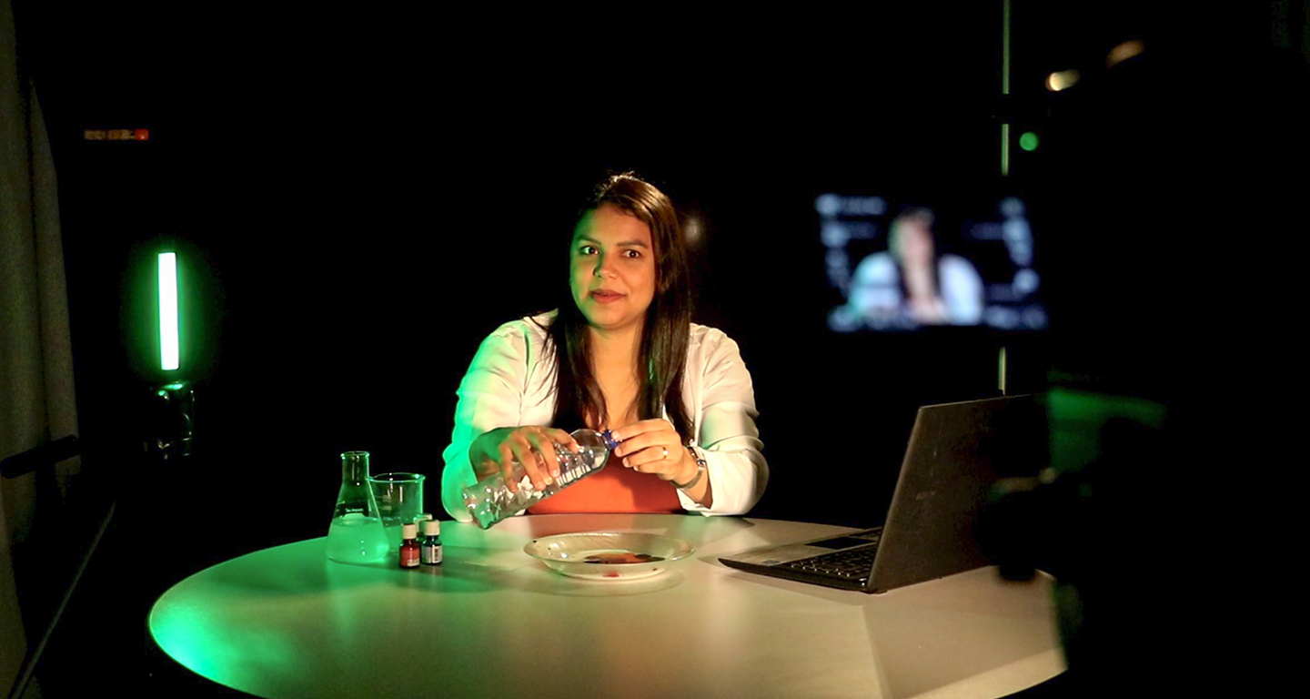 Uma mulher aparece sentada atrás de uma mesa redonda em um estúdio de gravação de videoaulas com iluminação verde ao fundo, demonstrando um experimento prático ao segurar uma garrafa plástica sobre um prato. À sua direita está um notebook aberto e, à esquerda, frascos de laboratório béquer e erlenmeyer, enquanto a silhueta de uma câmera de filmagem em primeiro plano e um monitor ao fundo contextualizam a produção de conteúdo educacional EAD da Unoeste.