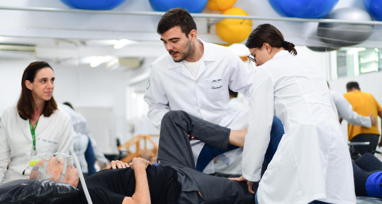 Estudantes de Fisioterapia realizam atendimento supervisionado na área de neurologia, auxiliando na reabilitação de paciente idoso.