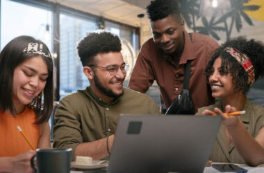 Quatro jovens adultos sentados à mesa, utilizando um notebook e conversando durante atividade em grupo em ambiente interno.