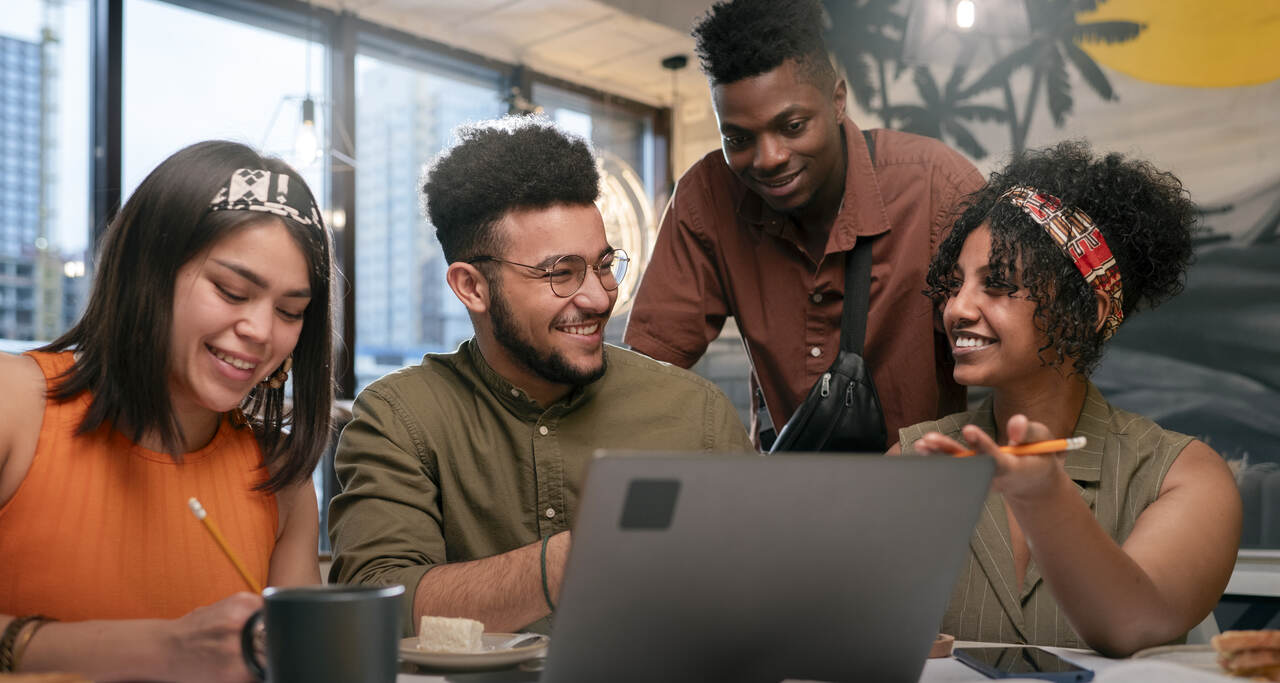 Quatro jovens adultos sentados à mesa, utilizando um notebook e conversando durante atividade em grupo em ambiente interno.