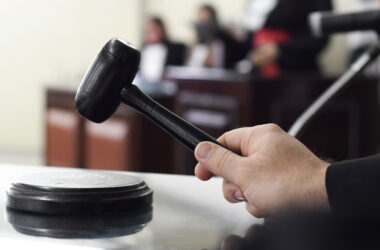 Mão segurando martelo de juiz sobre a mesa em um tribunal, representando carreiras e concursos na área do Direito.