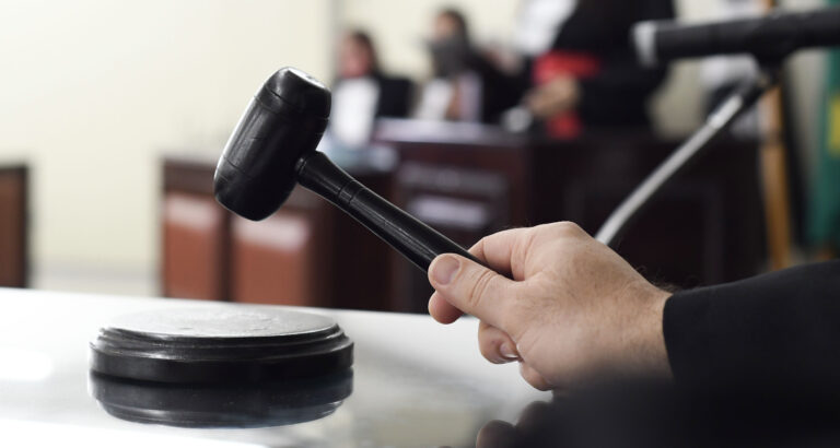 Mão segurando martelo de juiz sobre a mesa em um tribunal, representando carreiras e concursos na área do Direito.
