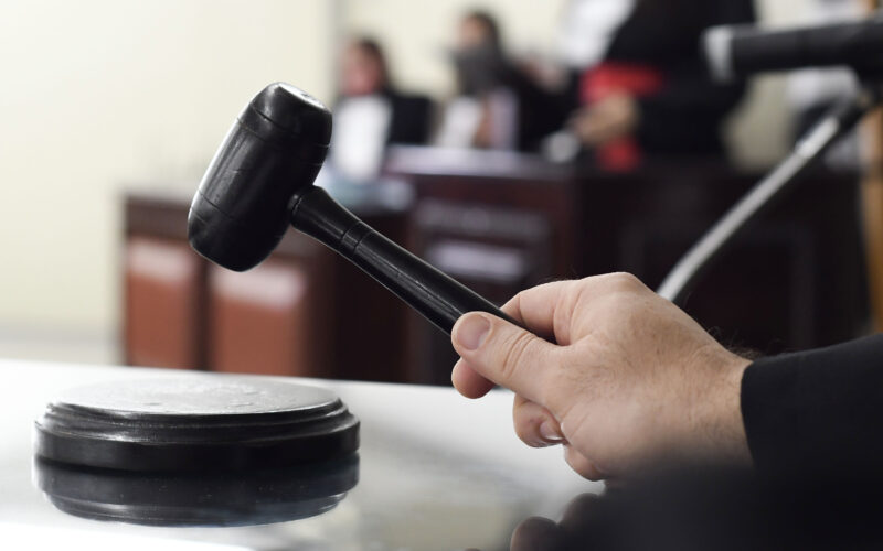 Mão segurando martelo de juiz sobre a mesa em um tribunal, representando carreiras e concursos na área do Direito.