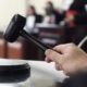 Mão segurando martelo de juiz sobre a mesa em um tribunal, representando carreiras e concursos na área do Direito.