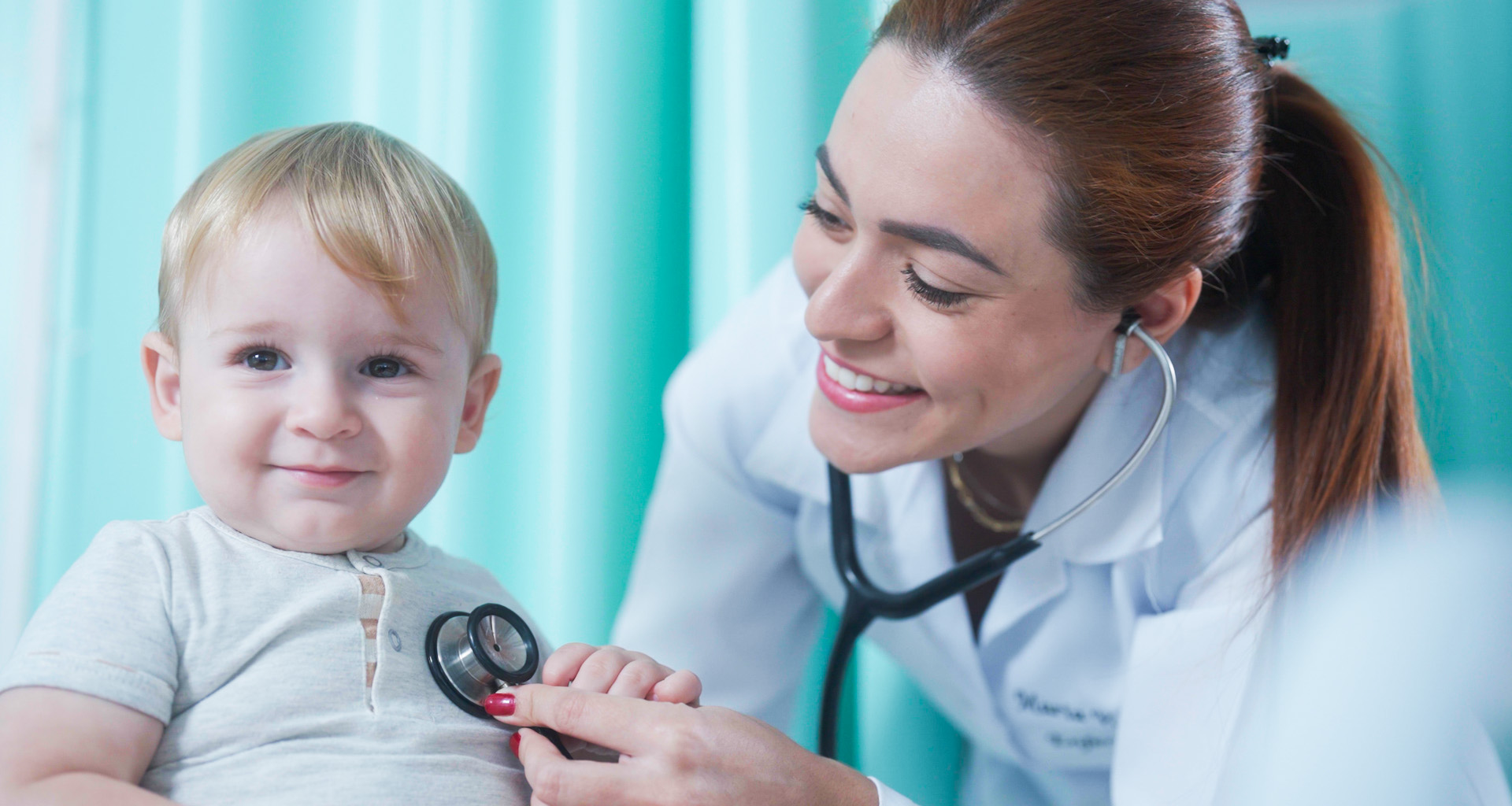Médica com estetoscópio sorrindo auscultando o coração de um menino de cabelos loiros, camiseta branca e sorrindo.