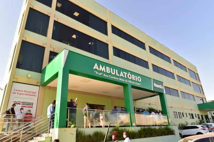 Fachada do Ambulatório Médico da Unoeste em um dia ensolarado. O prédio de vários andares, com tons de bege e janelas escuras, tem uma grande placa verde com a palavra "Ambulatório Profª Ana Cardoso Maia de Oliveira Lima" em branco. Algumas pessoas podem ser vistas na entrada e na varanda do prédio. Há também carros estacionados em frente.