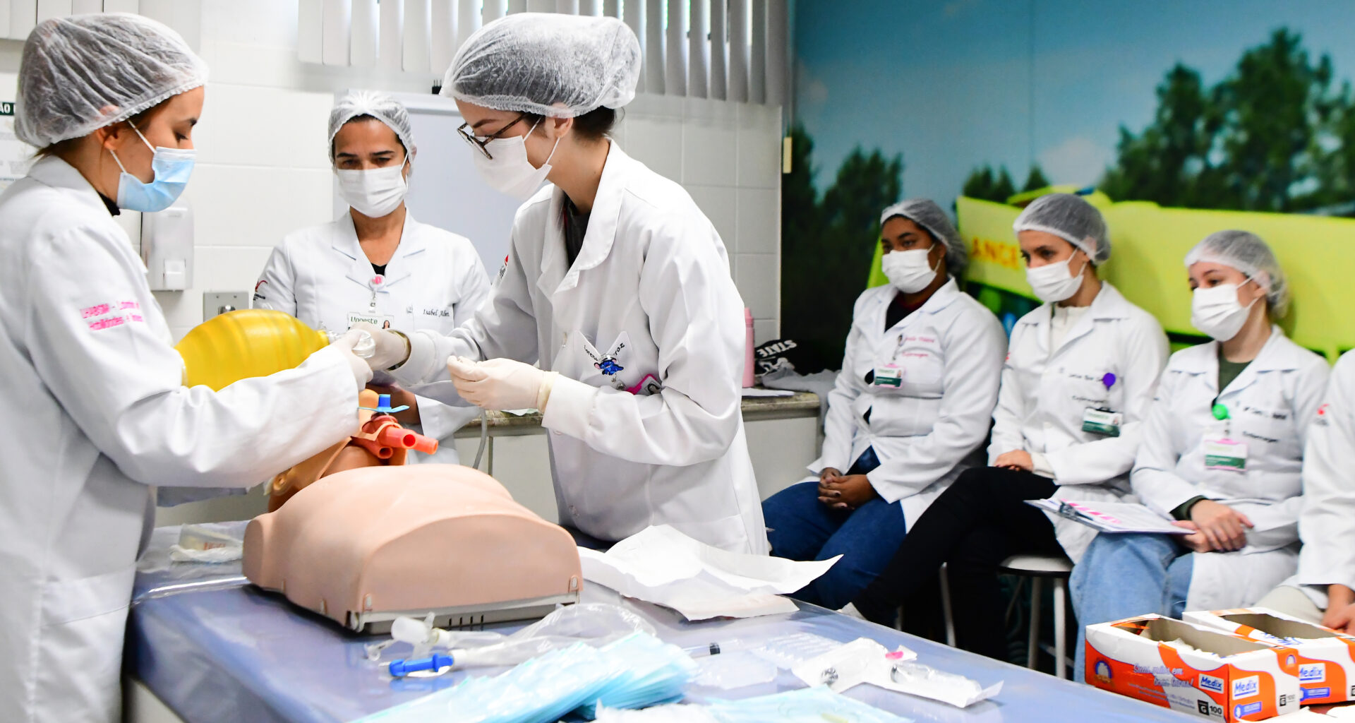 Estudantes do curso de Enfermagem da Unoeste realizam simulação prática com manequim em laboratório, enquanto colegas acompanham a atividade técnica em sala equipada.