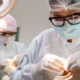 Um estudante de odontologia usando óculos de proteção, máscara, touca e luvas trabalha em um modelo de cabeça e pescoço. Um foco de luz está posicionado acima da cabeça do modelo. Ao fundo, outro aluno está trabalhando em outro modelo.