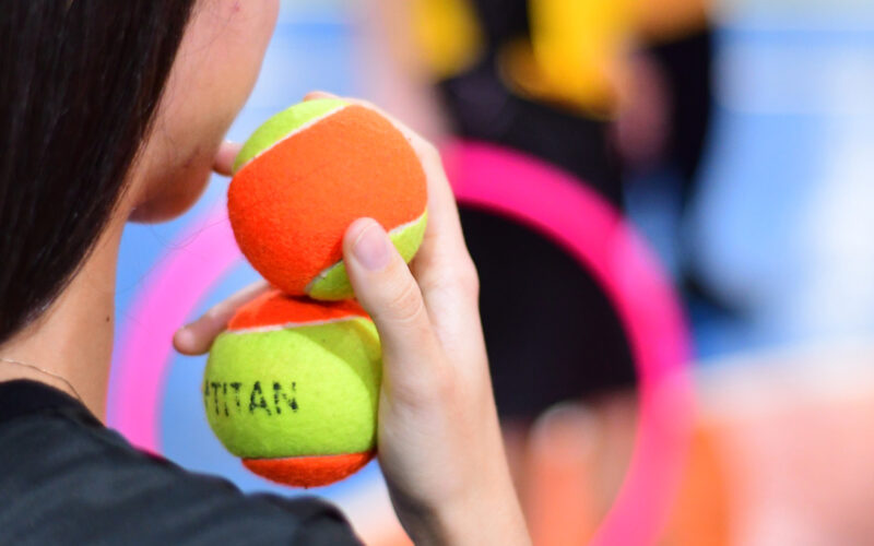 Estudante segurando duas bolas de tênis coloridas em ambiente esportivo, com outros objetos de atividade física ao fundo.