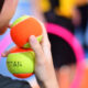Estudante segurando duas bolas de tênis coloridas em ambiente esportivo, com outros objetos de atividade física ao fundo.