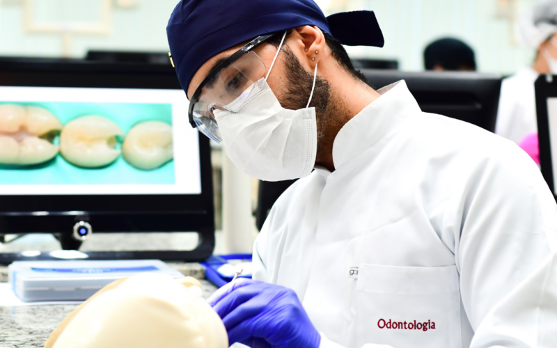 Estudante de odontologia em laboratório, usando jaleco, máscara e óculos de proteção enquanto realiza um procedimento prático em um modelo odontológico, com telas ao fundo exibindo imagens ampliadas de dentes. Representação do ensino prático em faculdade de odontologia.