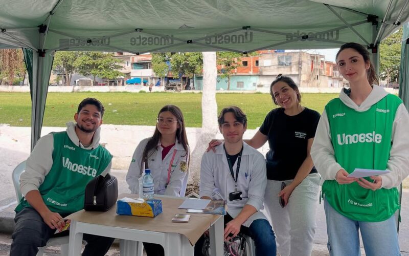 Estudantes universitários da Unoeste, em jalecos e coletes verdes, participam ativamente de um evento de extensão universitária ao ar livre. A imagem destaca o engajamento e a colaboração de alunos de ligas acadêmicas em iniciativas de voluntariado, exemplificando o aprendizado prático e a comunidade acadêmica.