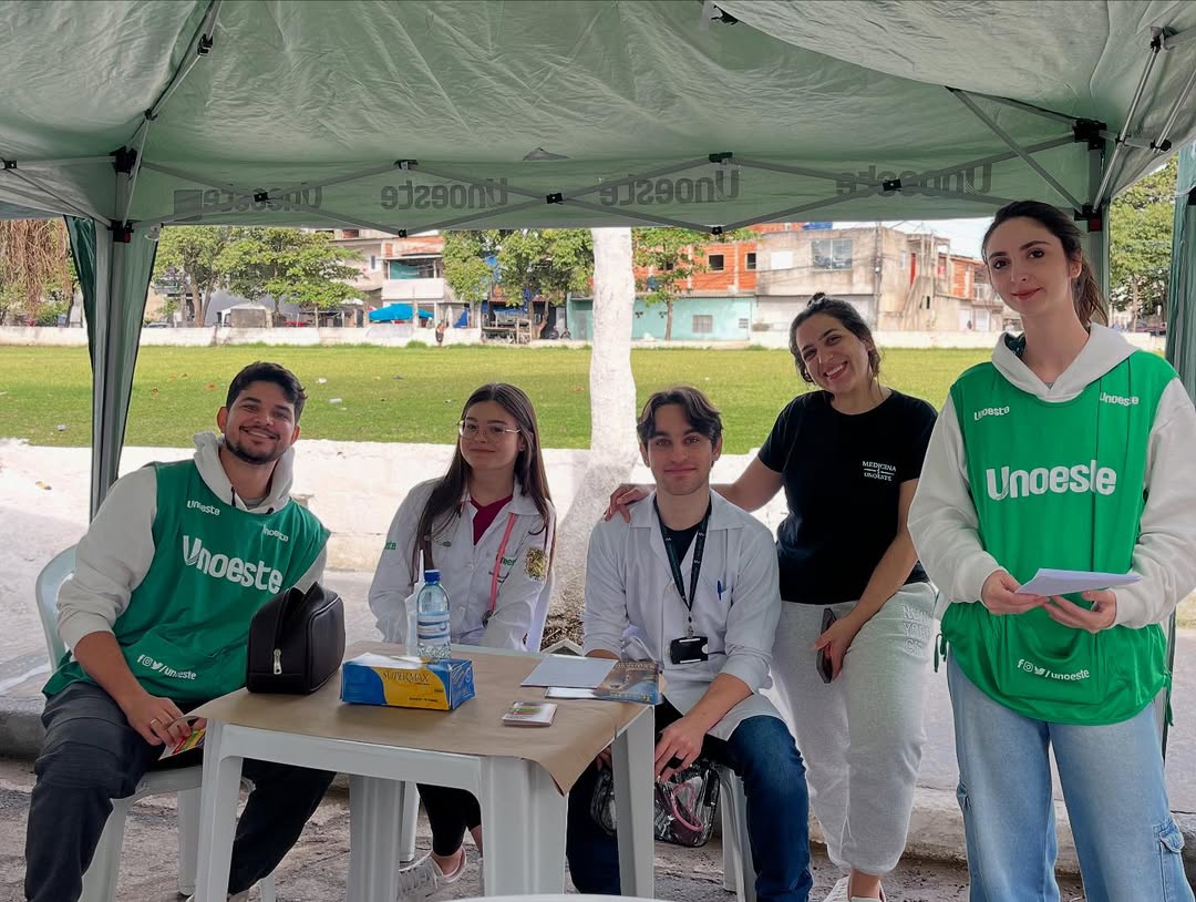 Estudantes universitários da Unoeste, em jalecos e coletes verdes, participam ativamente de um evento de extensão universitária ao ar livre. A imagem destaca o engajamento e a colaboração de alunos de ligas acadêmicas em iniciativas de voluntariado, exemplificando o aprendizado prático e a comunidade acadêmica.