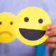 Duas expressões faciais em formato de círculo amarelo, uma sorridente e outra triste, sendo seguradas por uma pessoa. A imagem simboliza a dualidade das emoções e a importância da saúde emocional.