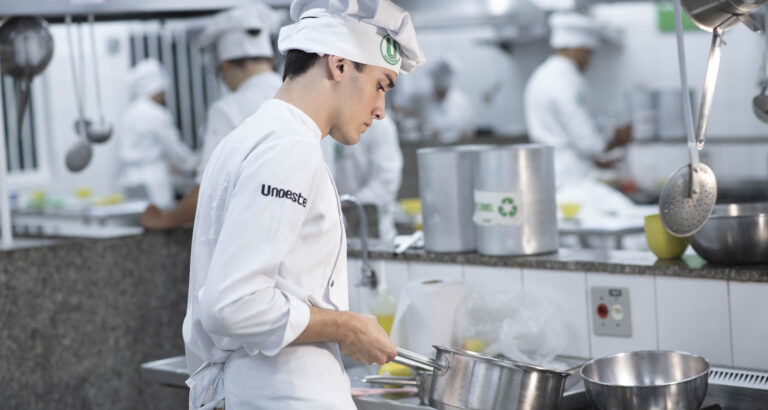 Um estudante do curso de Gastronomia da Unoeste trabalha concentrado na cozinha experimental da universidade. Ele veste um uniforme branco com a logo da instituição e um chapéu de chef, enquanto segura uma panela sobre o fogão. Ao fundo, outros alunos também estão em atividade, preparando receitas e utilizando utensílios de cozinha. O ambiente é profissional e bem equipado, com bancadas de inox e diversos instrumentos culinários.