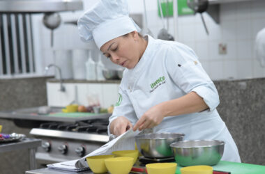 Estudante de Gastronomia da Unoeste realiza atividade prática em cozinha pedagógica, seguindo uma receita e preparando ingredientes em bancada equipada com utensílios e equipamentos profissionais.