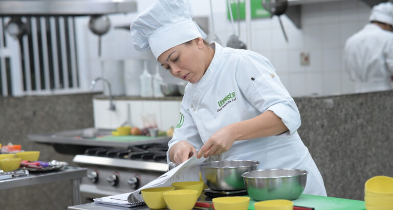 Estudante de Gastronomia da Unoeste realiza atividade prática em cozinha pedagógica, seguindo uma receita e preparando ingredientes em bancada equipada com utensílios e equipamentos profissionais.