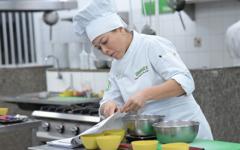 Estudante de Gastronomia da Unoeste realiza atividade prática em cozinha pedagógica, seguindo uma receita e preparando ingredientes em bancada equipada com utensílios e equipamentos profissionais.