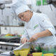 Estudante de Gastronomia da Unoeste realiza atividade prática em cozinha pedagógica, seguindo uma receita e preparando ingredientes em bancada equipada com utensílios e equipamentos profissionais.