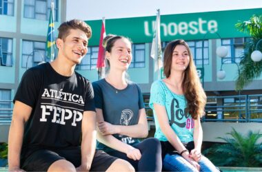 Três estudantes sentados em um banco no campus da Unoeste, com a fachada do prédio ao fundo exibindo o logotipo da universidade. Ao lado, bandeiras do Brasil e de São Paulo estão hasteadas, complementando o cenário acadêmico. O ambiente é cercado por vegetação, incluindo palmeiras e arbustos bem cuidados, transmitindo uma atmosfera acolhedora e organizada.