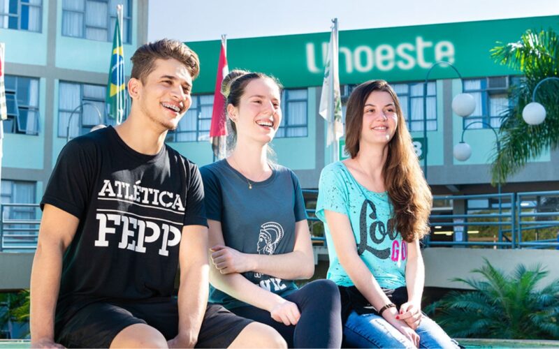 Três estudantes sentados em um banco no campus da Unoeste, com a fachada do prédio ao fundo exibindo o logotipo da universidade. Ao lado, bandeiras do Brasil e de São Paulo estão hasteadas, complementando o cenário acadêmico. O ambiente é cercado por vegetação, incluindo palmeiras e arbustos bem cuidados, transmitindo uma atmosfera acolhedora e organizada.