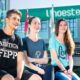 Três estudantes sentados em um banco no campus da Unoeste, com a fachada do prédio ao fundo exibindo o logotipo da universidade. Ao lado, bandeiras do Brasil e de São Paulo estão hasteadas, complementando o cenário acadêmico. O ambiente é cercado por vegetação, incluindo palmeiras e arbustos bem cuidados, transmitindo uma atmosfera acolhedora e organizada.