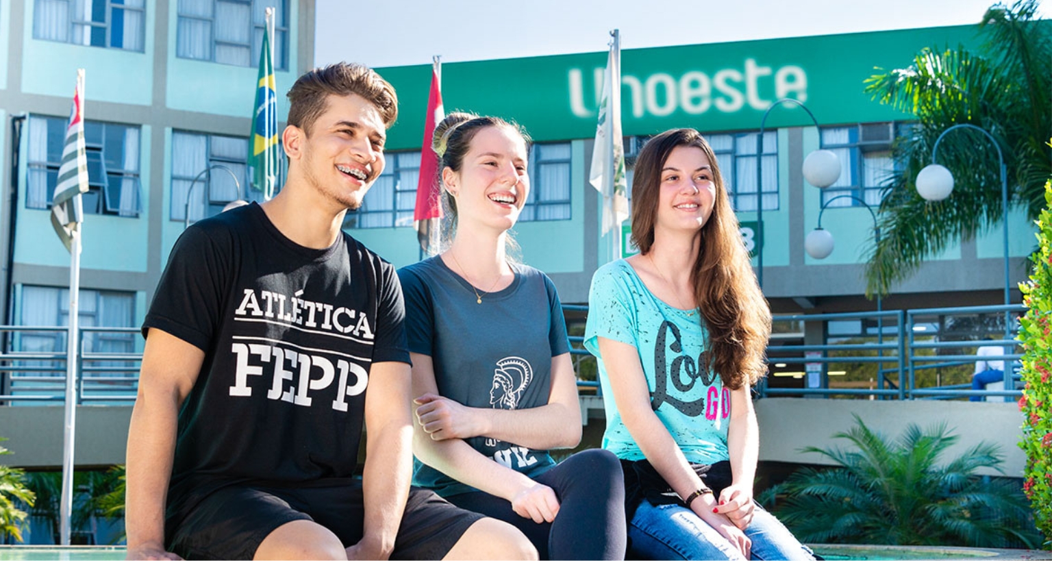 Três estudantes sentados em um banco no campus da Unoeste, com a fachada do prédio ao fundo exibindo o logotipo da universidade. Ao lado, bandeiras do Brasil e de São Paulo estão hasteadas, complementando o cenário acadêmico. O ambiente é cercado por vegetação, incluindo palmeiras e arbustos bem cuidados, transmitindo uma atmosfera acolhedora e organizada.