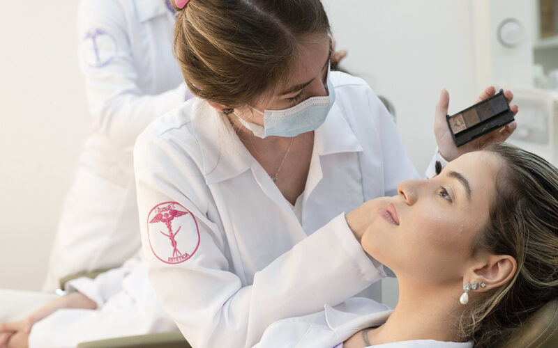 Estudante de Estética e Cosmética realizando procedimento de maquiagem facial em paciente durante atividade prática supervisionada, destacando técnicas profissionais de cuidado com a pele e embelezamento.