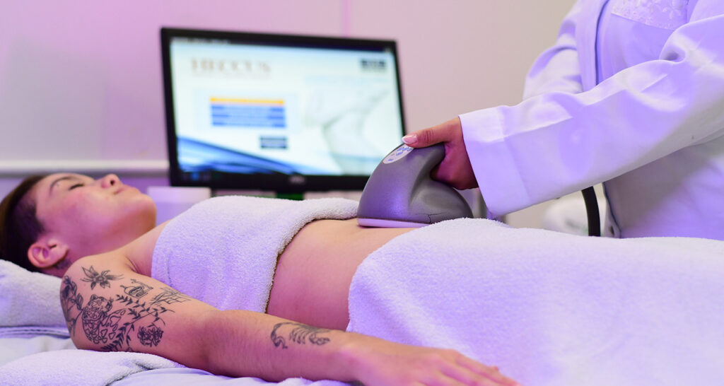 Profissional de Estética realizando procedimento corporal com equipamento de alta tecnologia na região abdominal de uma paciente, destacando práticas modernas utilizadas no curso de Estética e Cosmética.