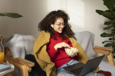 Mulher jovem sorridente, usando fones de ouvido e notebook no colo, comendo pipoca em uma poltrona confortável. Cena ideal para assistir a filmes sobre inteligência artificial em casa, desfrutando de entretenimento digital e o fascinante universo da IA.