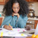 Como trabalhar e estudar? Na foto, uma jovem de pele negra sentada com um notebook e várias folhas de caderno espalhadas estudando.