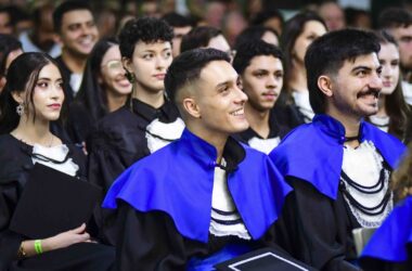 Uma imagem vibrante da colação de grau na Unoeste, capturando a alegria e o alívio de vários formandos. Em primeiro plano, três jovens sorriem, com os olhos fixos em algo fora do quadro, irradiando felicidade. O homem à direita tem um sorriso largo e um bigode, enquanto o do centro, com um corte de cabelo moderno, também sorri amplamente. À esquerda, uma mulher com longos cabelos escuros e brincos elegantes exibe um sorriso mais contido, mas igualmente satisfeito. Todos vestem togas pretas e capelos azuis, símbolos da conquista acadêmica. Ao fundo, outros formandos são visíveis, alguns sorrindo, outros com expressões de contemplação, todos compartilhando o mesmo momento de celebração.