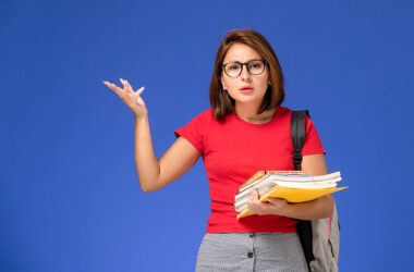 Vale a pena fazer faculdade? Na foto, uma jovem branca, de cabelo liso e curto na altura dos ombros, óculos de grau, com uma camiseta vermelha, uma calça cinza e segurando alguns livros com uma mochila nas costas.